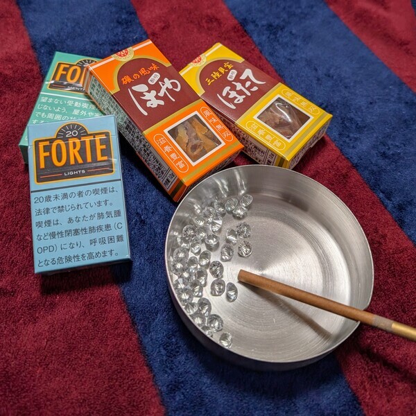 ほや酔明（橙色）とほたて酔明（黄色）を、本物のタバコ（青と緑のFORTE）と金属製の灰皿と一緒に並べた様子